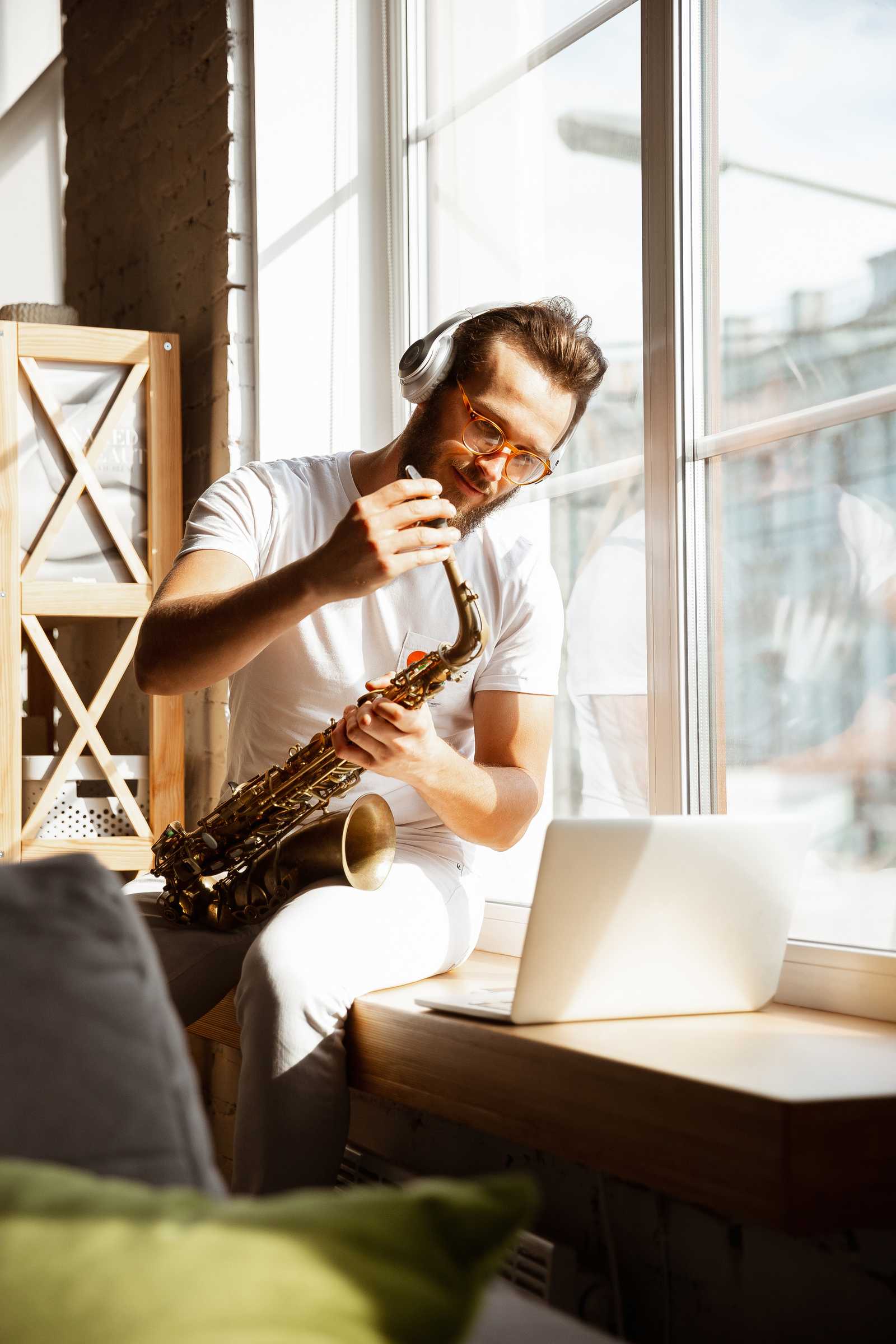 a man practicing saxophone by a window with a laptop